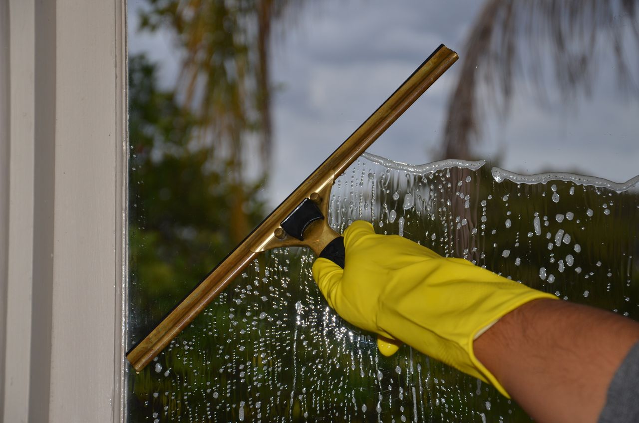 services-04 Close-up of hand cleaning window with squeegee outdoors using yellow gloves.